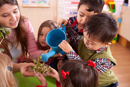 Educator holding a plant while young children water the plant with a watering can
