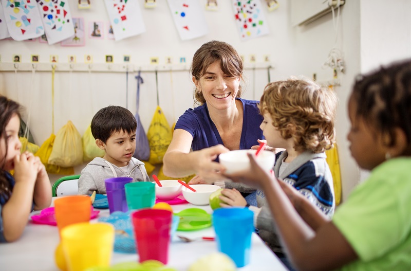 Educator and children sitting together at meal time using colourful utensils and dishes