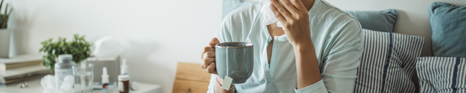 Woman sick in bed, blowing her nose, surrounded by tissues. Holding a hot drink in a mug.