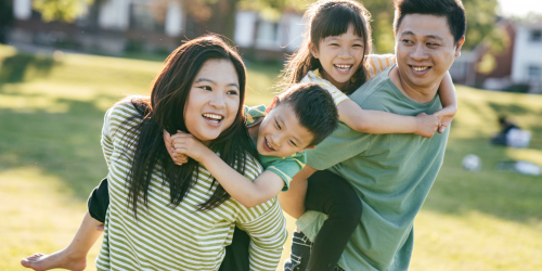 A family of four are in a park. Each of the parents is giving one child a piggy back.