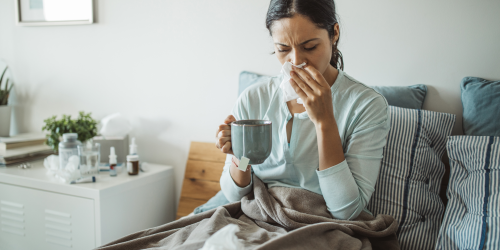 Woman sick in bed, blowing her nose, surrounded by tissues. Holding a hot drink in a mug.
