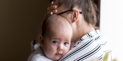 A woman can be seen holding a baby. The woman is faced away from the camera, the baby is looking into the camera.
