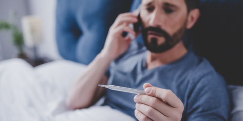 Man sick, sitting in bed, talking on the phone and holding a thermometer