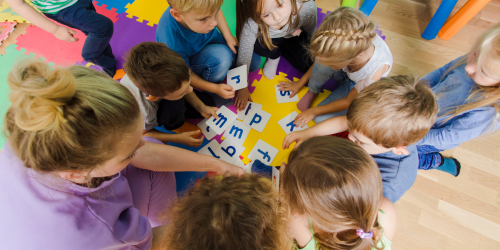 Photo is taken from above. An educator is playing with letter cards with a group of children sitting on the floor.