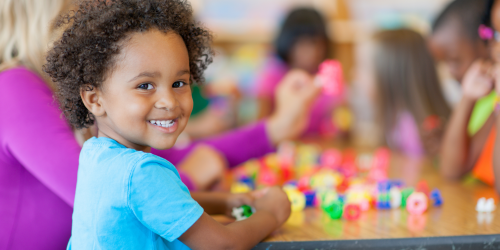 Child playing with blocks on table, looks back over their shoulder smiling at the camera.