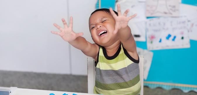 Young boy reaching hands out towards the camera, with a disruptive expression in classroom setting