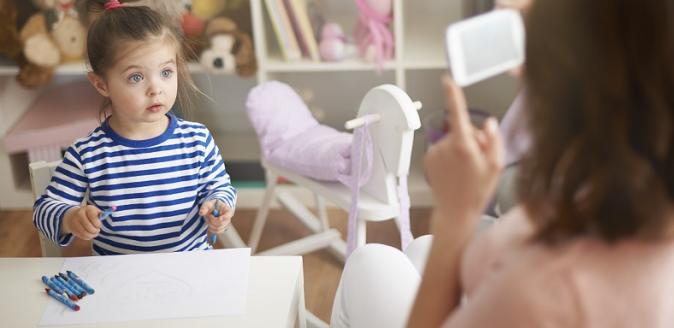 Woman holding up mobile device, capturing image of child sitting at table with paper and crayons