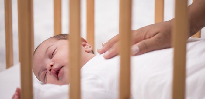 Newborn baby asleep in a cot with a caregiver's hand resting gently on the baby's torso