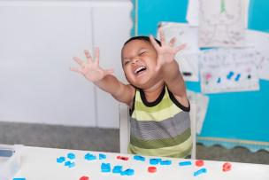 Young boy reaching hands out towards the camera, with a disruptive expression in classroom setting