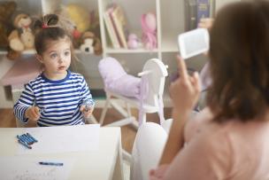 Woman holding up mobile device, capturing image of child sitting at table with paper and crayons