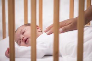 Newborn baby asleep in a cot with a caregiver's hand resting gently on the baby's torso